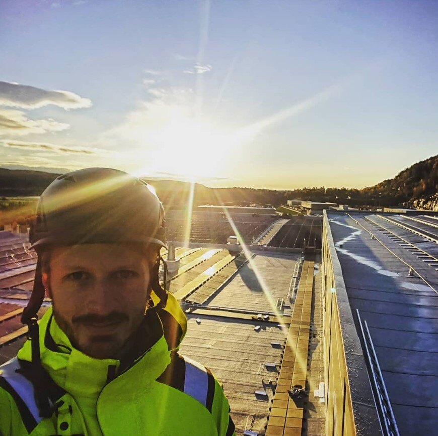 En person i gul refleksjakke og hjelm står på et tak med solcellepaneler, med solen lavt på himmelen i bakgrunnen som skaper et varmt lys og lange skygger.